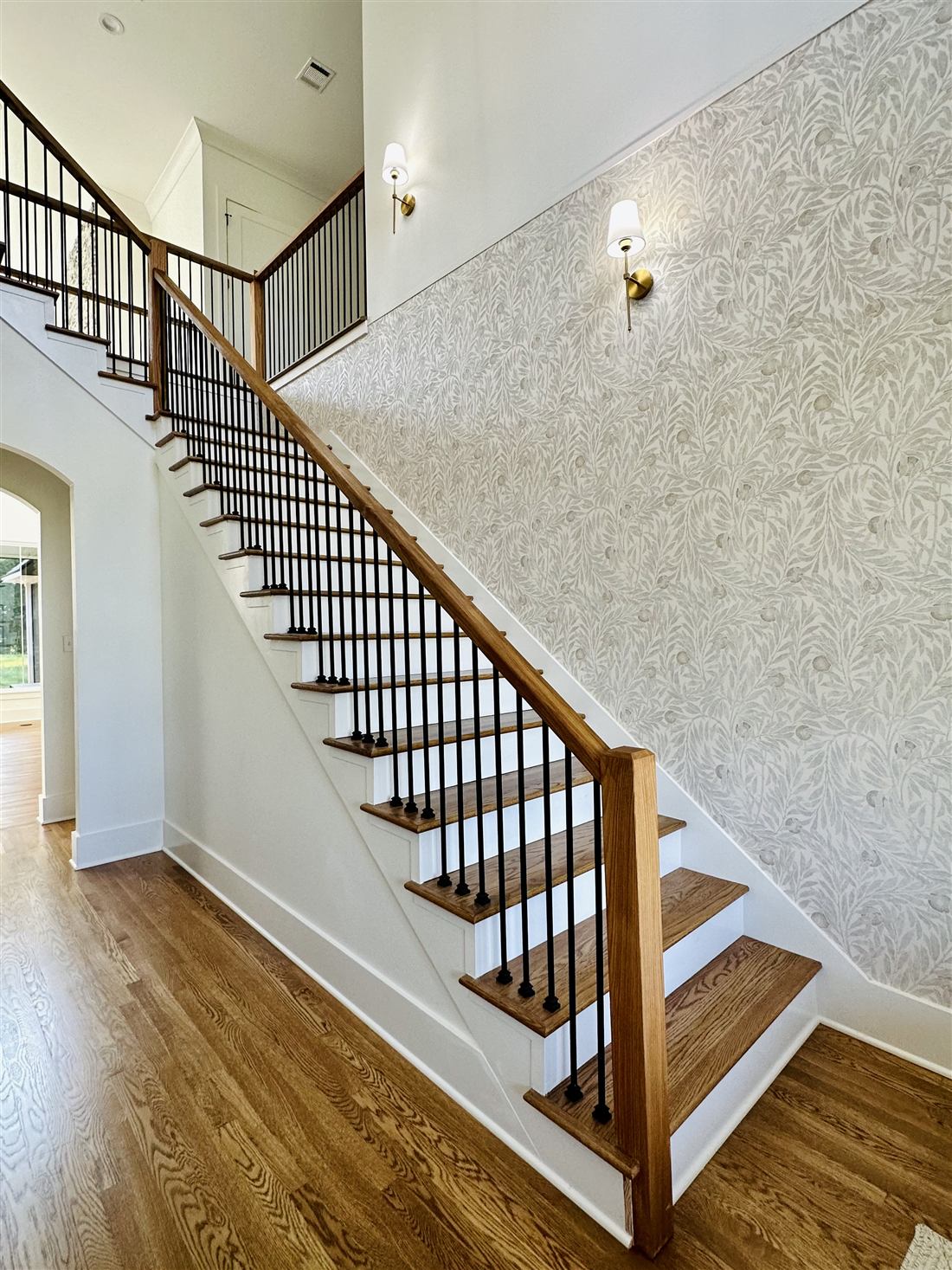 Custom oak staircase with iron balusters, brass sconces and wallpaper feature wall in a new custom home